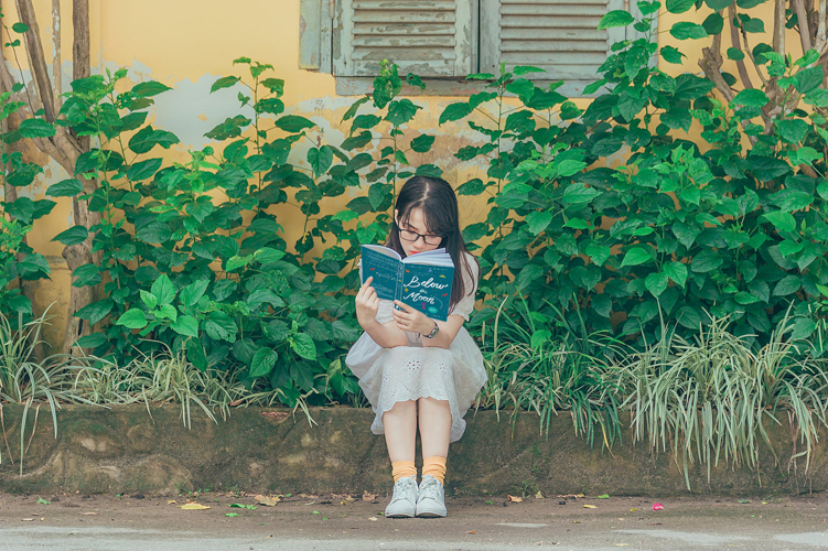 Girl in front of shrubbery reading Below the Moon by Alexis Marie Chute